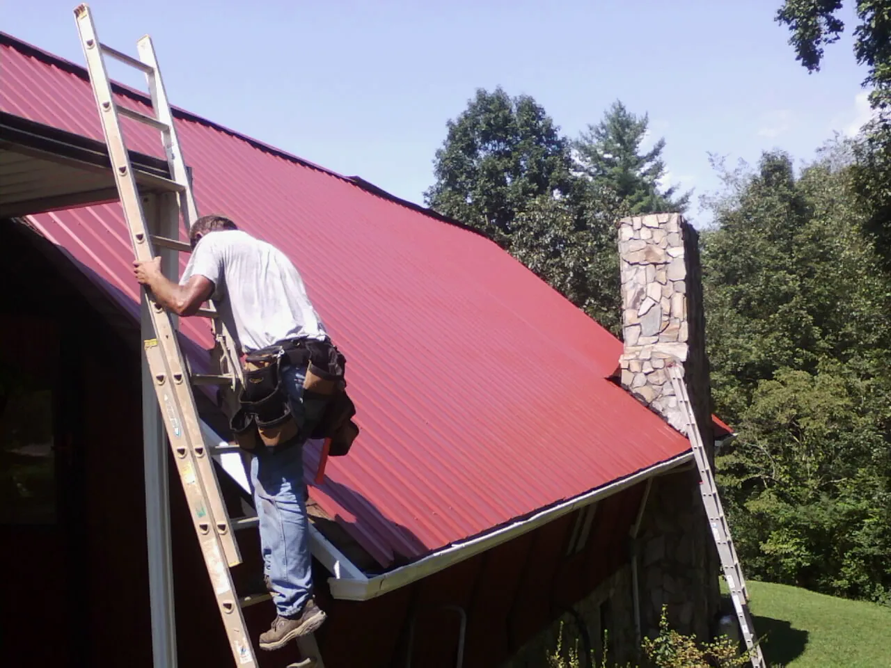 Roofer on a ladder beside a red metal roof and stone chimney in Richmond